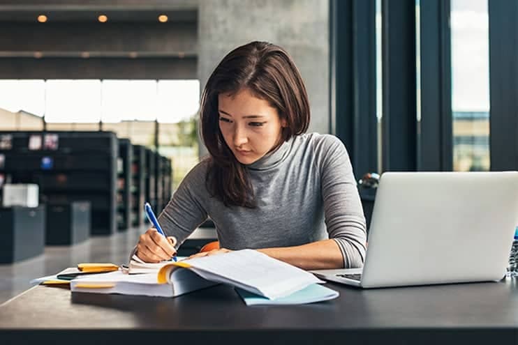 Young Woman Sitting In Library Studying With Book And Laptop